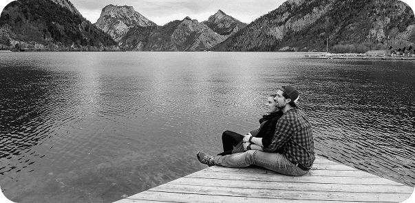 Two people sitting on wooden dock with mountains in background
